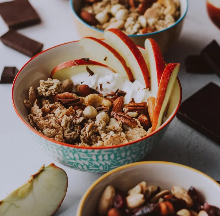 Em uma mesa há três tigelas com frutas e cereais dentro, sendo eles maçã, nozes e castanhas. Também há chocolate partido e espalhado pela mesa.