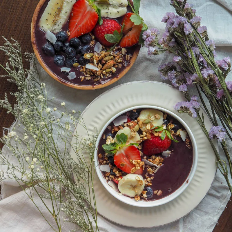 Imagem de uma mesa de madeira, com uma toalha branca. Possui duas tigelas de açaí, com morangos, banana, mirtilo e granola. Também há galhos de flores brancas e lilás ao lado das tigelas para decorar.